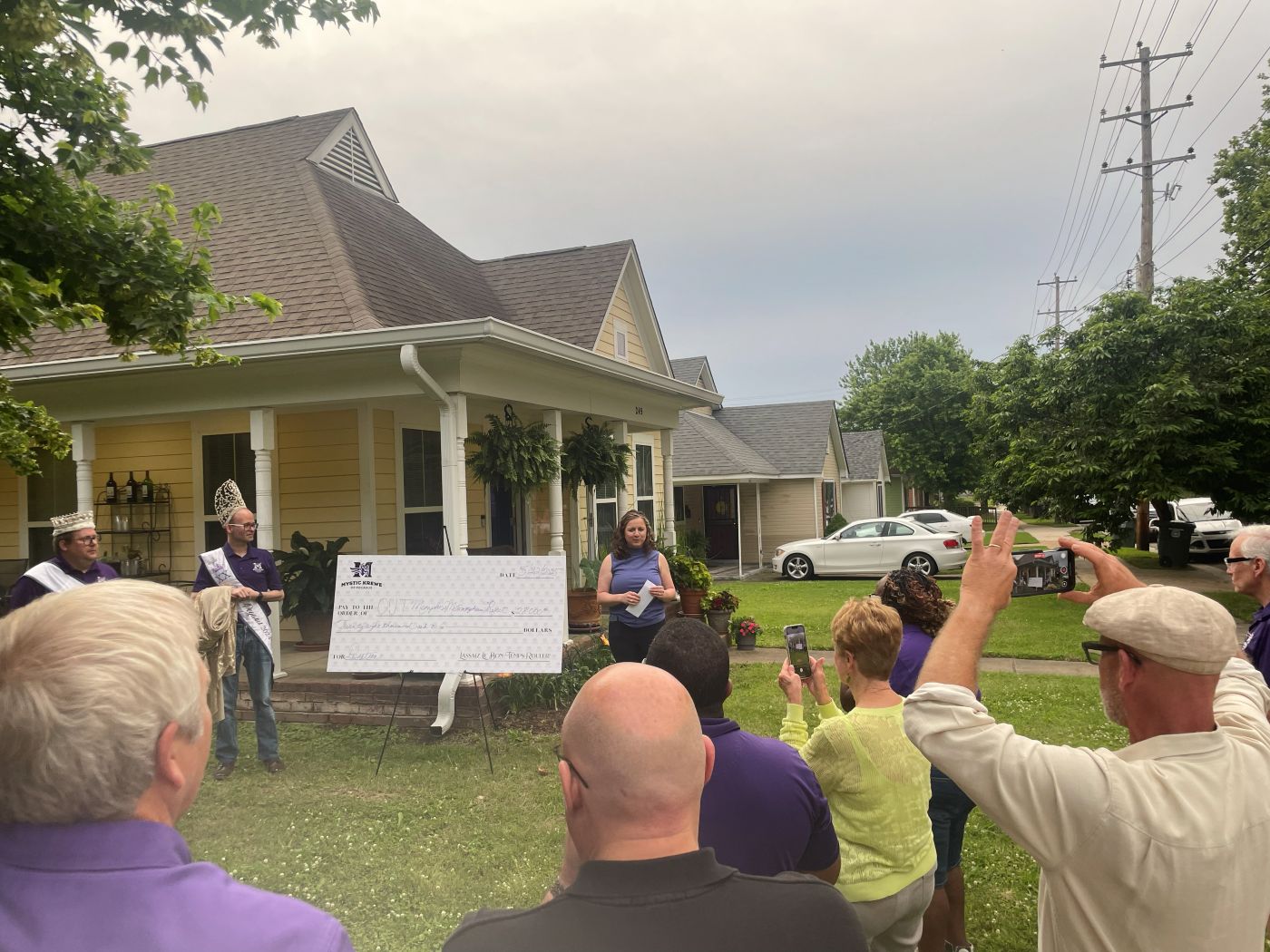 Krewe members in crowns and purple hold an oversized donation check on a lawn while a charity staff member speaks.