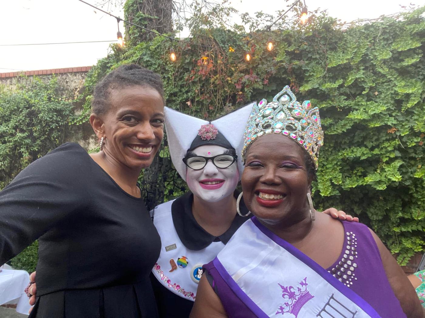 Three guests at the Krewe silent auction laughing together outdoors, one in a rhinestone crown and sash.
