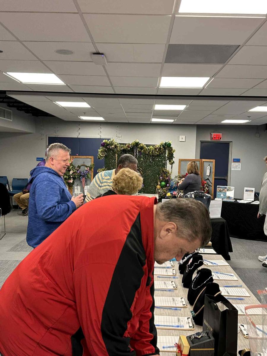 A guest in a red jacket leans over a row of clipboards, reading the bid sheets before writing.