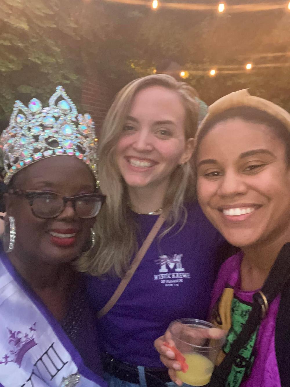 Three guests crowd into a selfie, one in a tall rhinestone crown and Krewe sash, drinks in hand.
